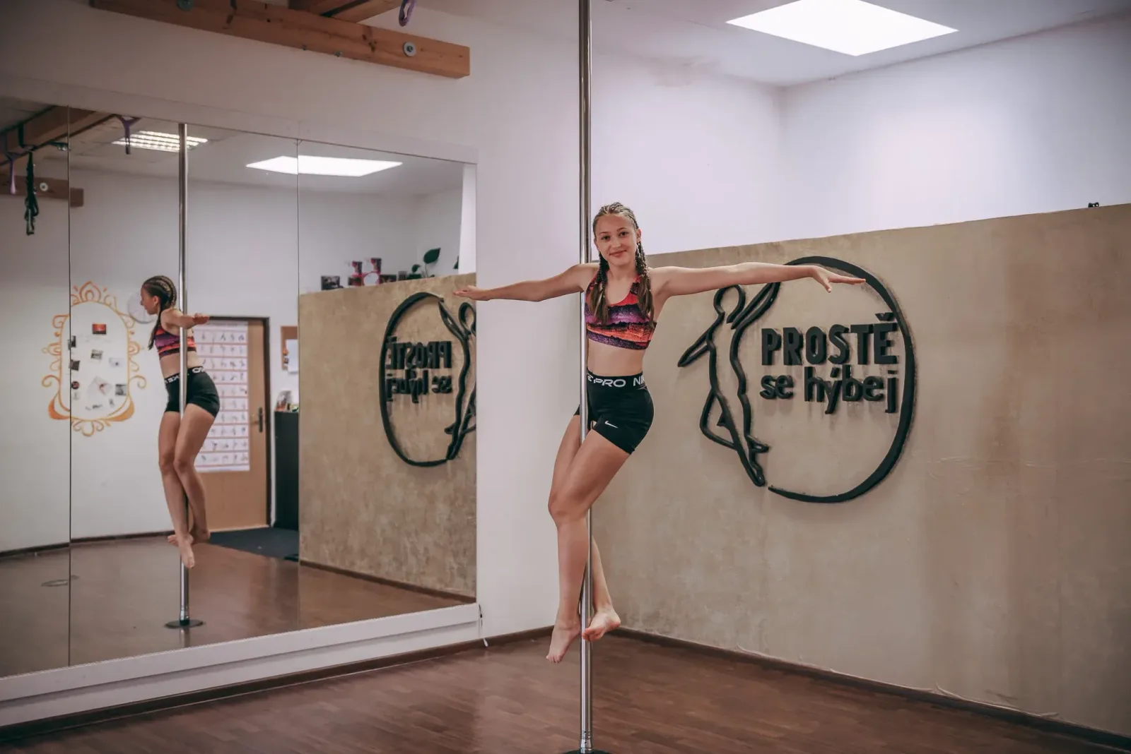 Pole dance – mladá žena balancuje na tyči s rozpaženýma rukama ve studiu Prostě se hýbej, Opava.